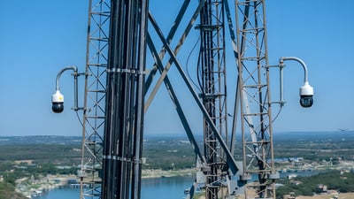 Two dome security cameras mounted on a metal utility tower overlooking a river and forested landscape.