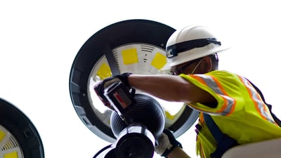 A technician in a hard hat and safety vest installs a circular LED street lighting fixture against a bright sky.