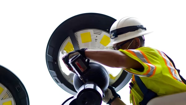 A technician in a hard hat and safety vest installs a circular LED street lighting fixture against a bright sky.