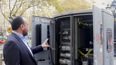 A man in a suit gestures toward the internal wiring and hardware of an open street-side electrical cabinet in Barcelona.