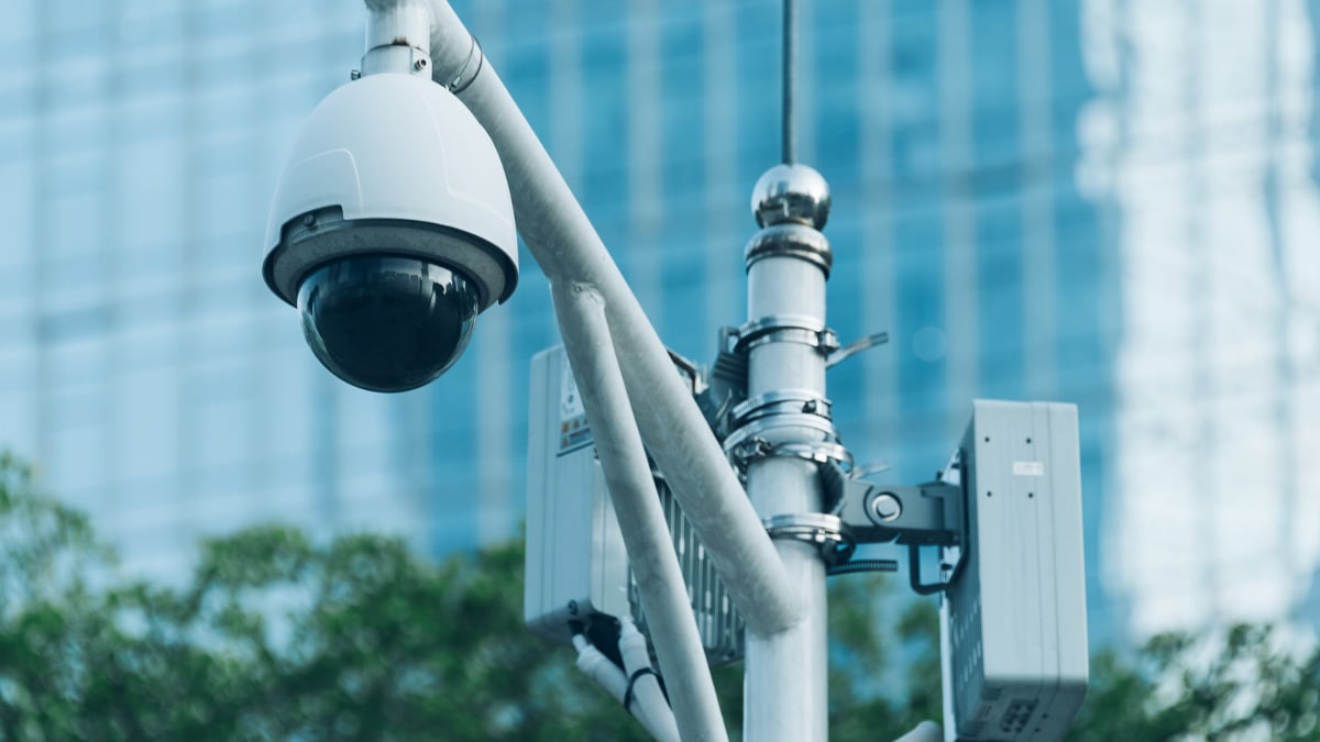 A dome security camera and telecommunications equipment mounted on a metal pole with a blue glass building behind it