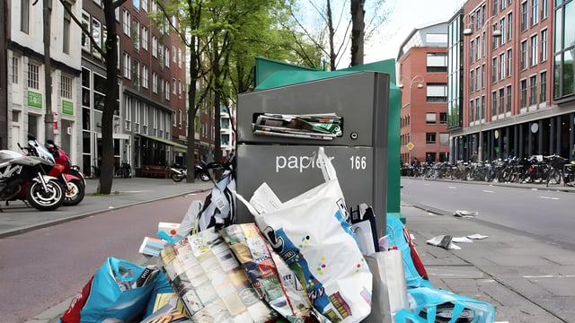Overflowing paper recycling bin labeled "papier" on a city street in Amsterdam with loose trash on the sidewalk.