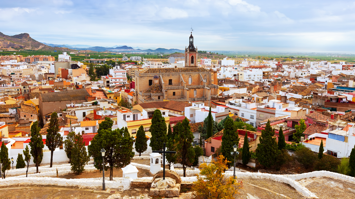 Elevated view of a historic Spanish town with a central stone church, terracotta-roofed buildings, and distant hills