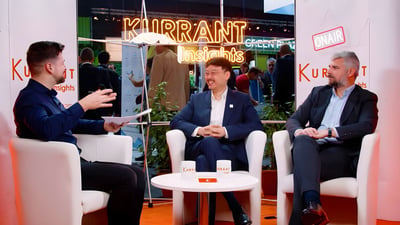 Three men in suits sit in white armchairs for a Kurrant Insights interview at a trade show booth with neon signage.