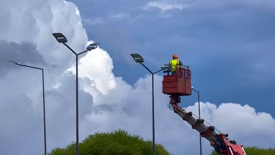 A technician in a cherry picker bucket repairs a LED street light against a cloudy sky.