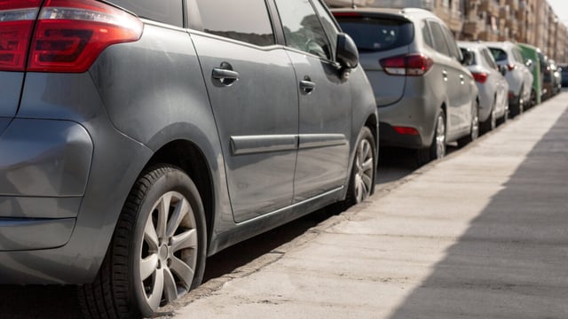 A row of cars parked bumper to bumper along a sunlit city street kerb, with apartment buildings in the background