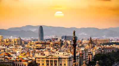 Barcelona skyline at sunset featuring the Torre Glòries and Columbus Monument against a backdrop of mountains.