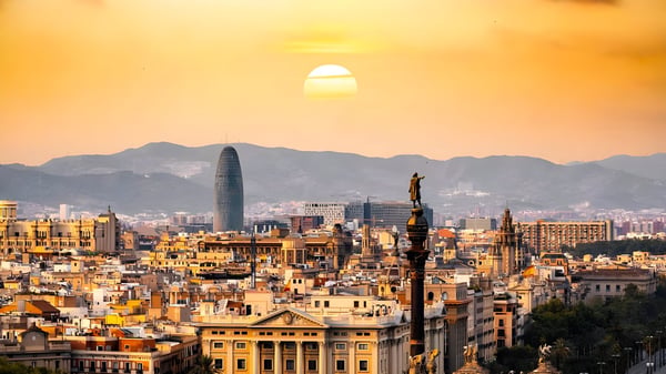 Barcelona skyline at sunset featuring the Torre Glòries and Columbus Monument against a backdrop of mountains.