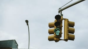 A yellow traffic light with a green signal illuminated, hanging from an overhead arm against a cloudy sky.