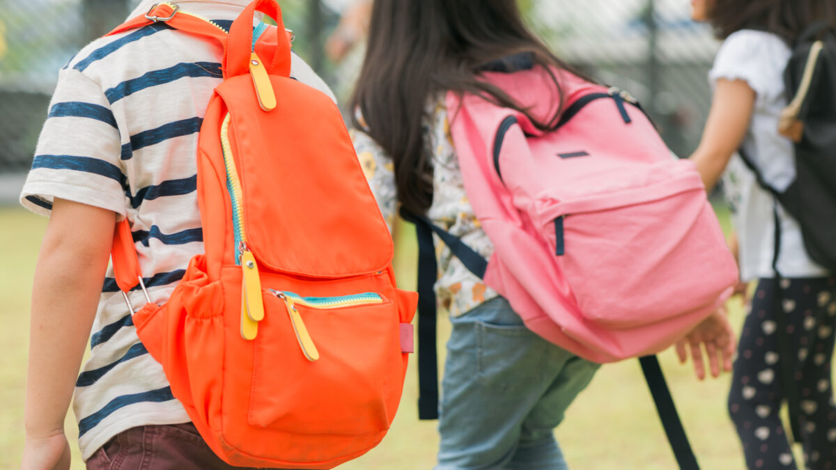 Three children with backpacks, one orange and one pink, walk away from the camera in an outdoor setting.