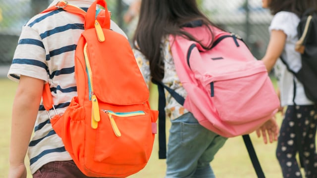 Three children with backpacks, one orange and one pink, walk away from the camera in an outdoor setting.