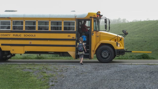 A child with a backpack boards a yellow county public schools bus on a rural road with its extended stop arm visible at the front