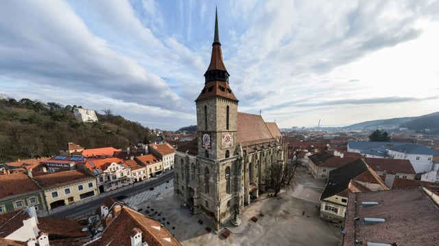 Wide aerial view of the Black Church tower and surrounding terracotta rooftops of old Brașov under a cloudy sky with hills beyond