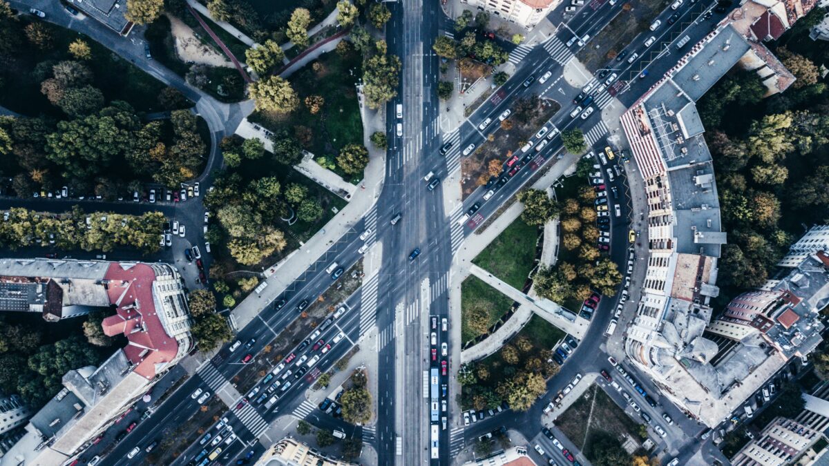 Aerial view of a city street intersection with multiple lanes of traffic, cars, buses, surrounding buildings, and green spaces.