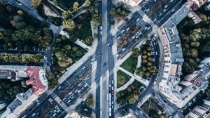 Aerial view of a city street intersection with multiple lanes of traffic, cars, buses, surrounding buildings, and green spaces.