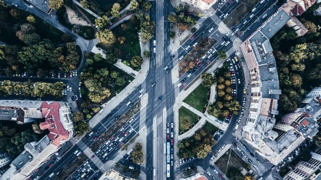 Aerial view of a city street intersection with multiple lanes of traffic, cars, buses, surrounding buildings, and green spaces.
