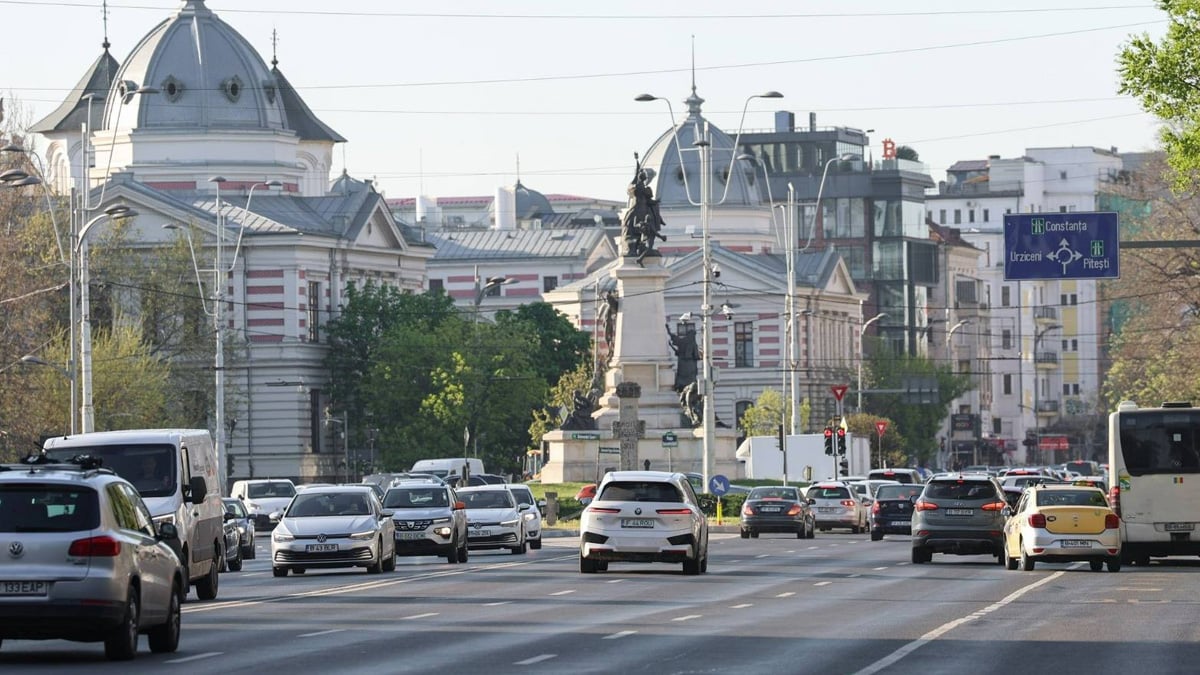 Heavy traffic on a multi-lane road in Bucharest near University Square with historic buildings and a blue overhead sign.