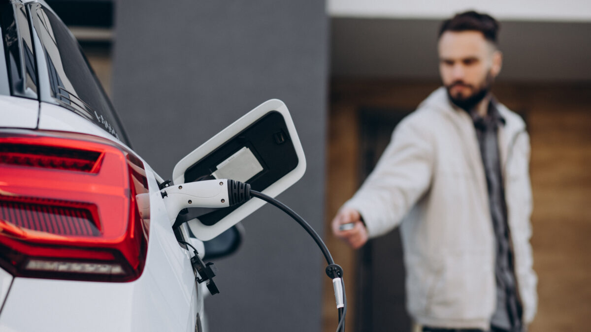 A man in a light jacket stands next to a white electric vehicle with a charging cable plugged into its rear port.