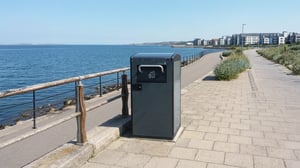 A solar-powered smart waste bin stands on a paved coastal promenade next to a metal railing and the sea.