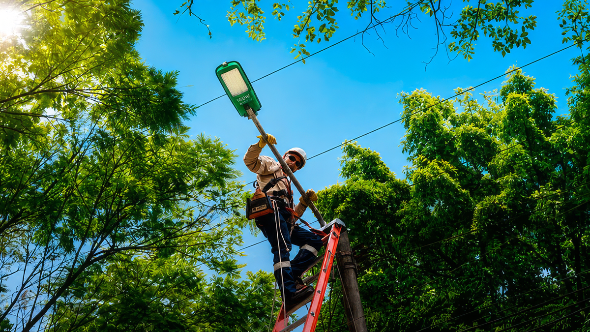 A technician on a ladder installs a green LED streetlight fixture on a utility pole surrounded by green trees.