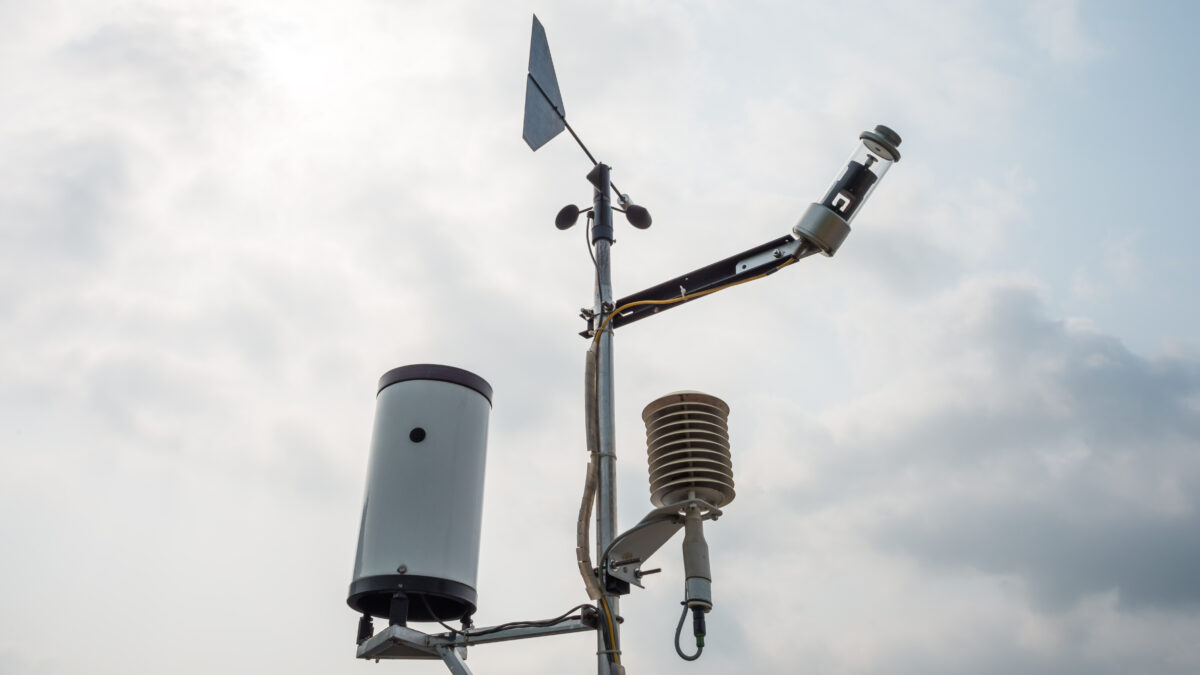 Weather station pole with an anemometer, wind vane, rain gauge, and radiation shield against a cloudy sky.