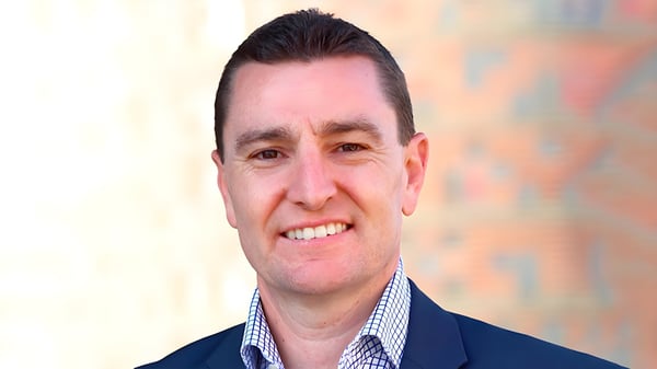 Headshot of a man wearing a blue blazer and a checkered button-down shirt against a blurred background.