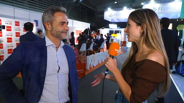 Joaquim Alvarez being interviewed by a woman with an orange microphone at a crowded indoor technology trade show.