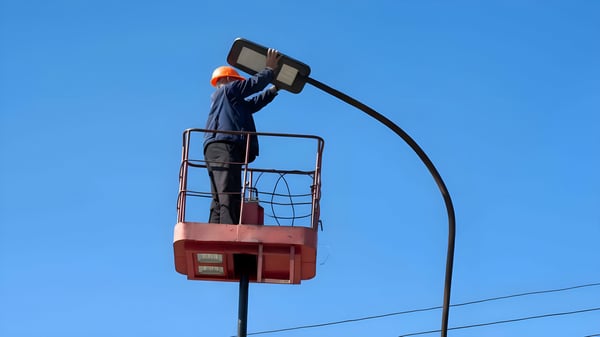 A utility worker in a bucket lift installs an LED street light fixture against a clear blue sky.