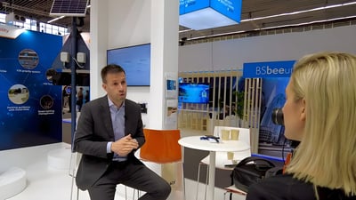 A man in a suit being interviewed by a woman at a technology trade show booth with a solar-powered pole in the background.