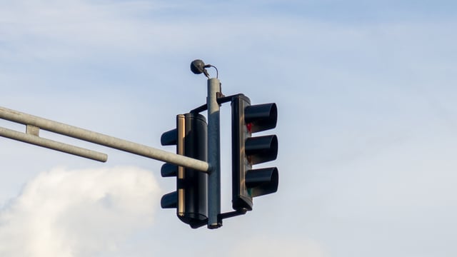 A traffic light with a small sensor mounted on top of the metal support arm against a blue sky with clouds.