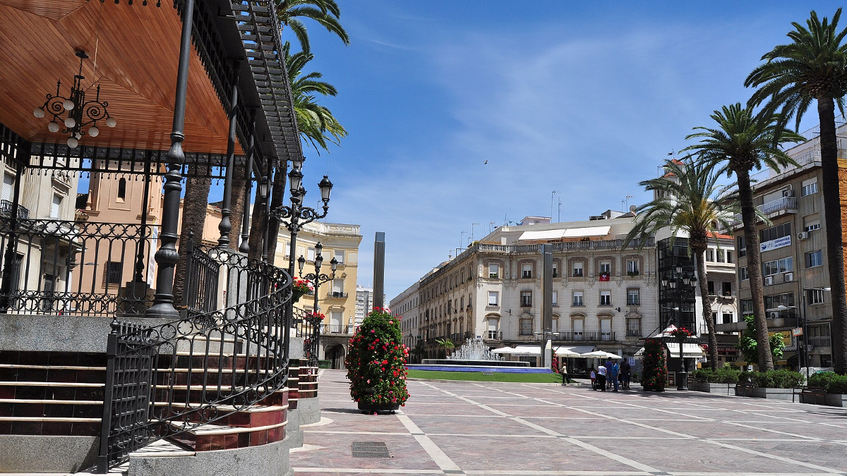 Sunny public plaza in Huelva featuring an ornate bandstand, street lights, palm trees, and surrounding buildings.