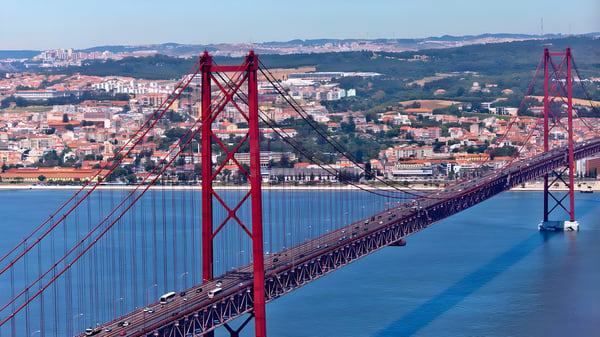 Aerial view of the red 25 de Abril Bridge spanning the Tagus River with the Lisbon cityscape in the background.