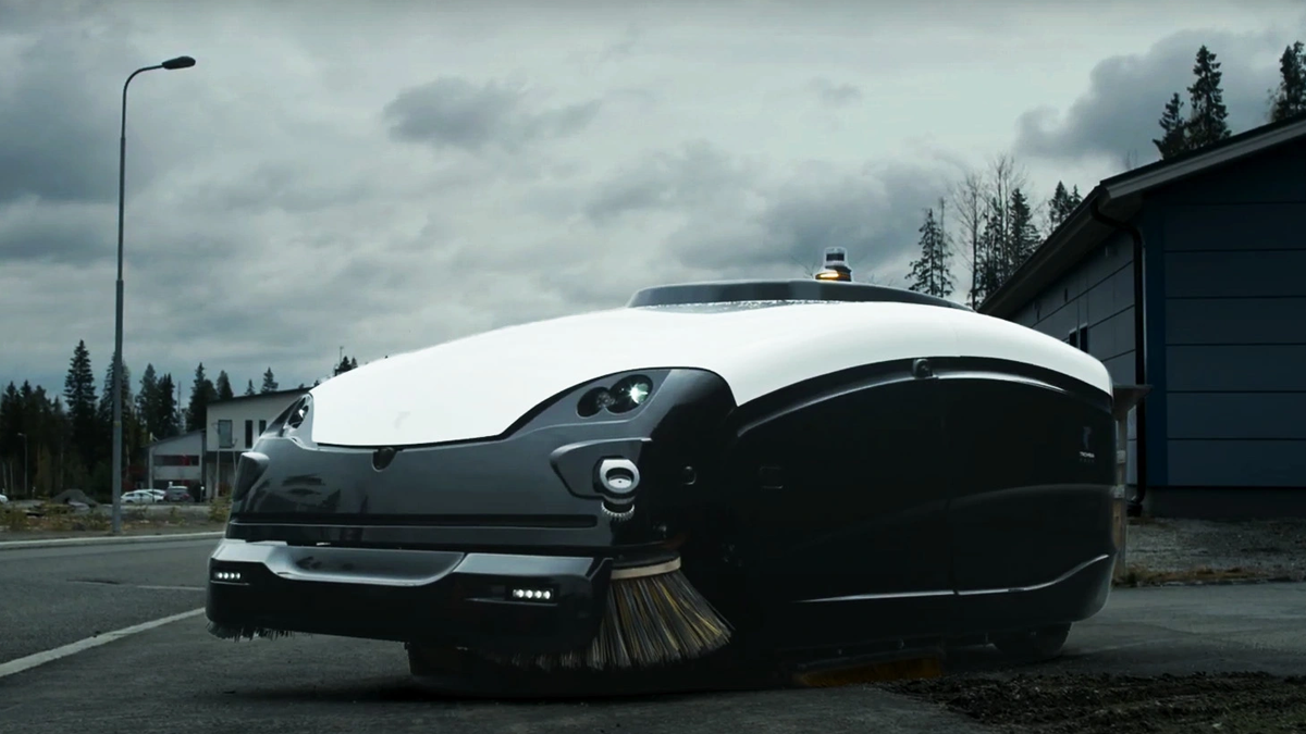 A Trombia Technologies autonomous electric street sweeper with sweeping brushes parked on asphalt under cloudy skies.