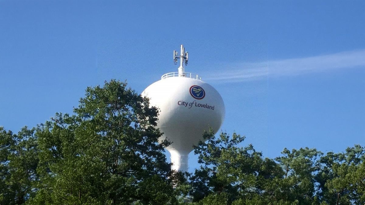 White City of Loveland water tower with telecommunications antennas rising above a green treeline under a clear blue sky.