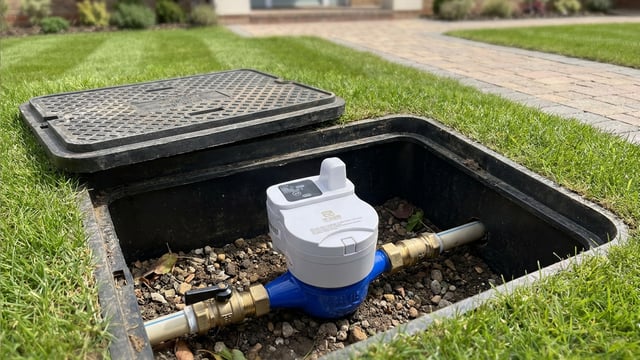 A white and blue smart water meter installed in an open underground utility box in a grassy lawn.
