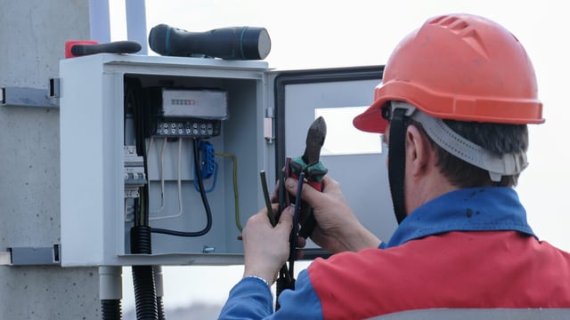 Electrician in an orange hard hat cuts wiring with pliers inside an open electricity meter box mounted on a concrete pole