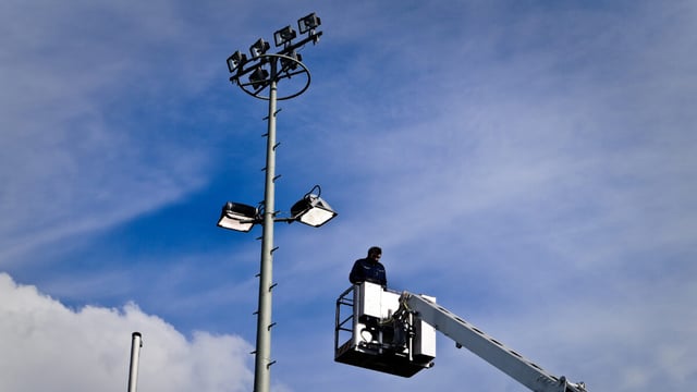 A worker in a hydraulic lift basket beside a tall metal pole with floodlight fixtures against a blue sky with scattered clouds