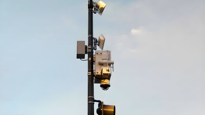 A black utility pole mounted with a gunshot detection sensor, dome security camera, and floodlights against a pale sky.