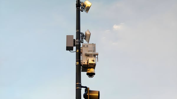 A black utility pole mounted with a gunshot detection sensor, dome security camera, and floodlights against a pale sky.