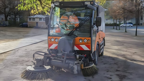 A compact orange and white street sweeper vehicle cleaning a paved public square in Basel.