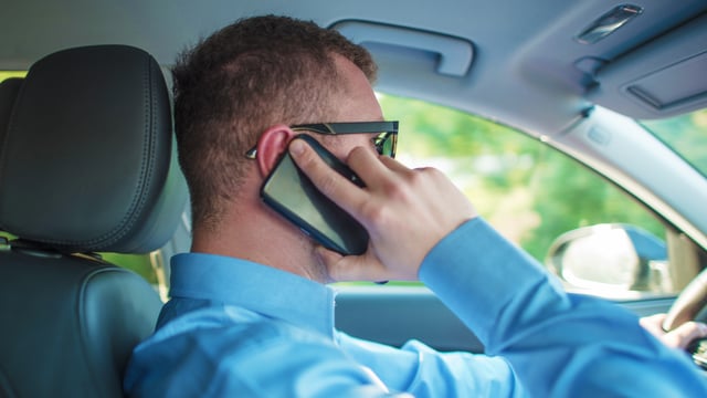 A man in a blue shirt talks on a smartphone while driving a car, seen from the passenger side.