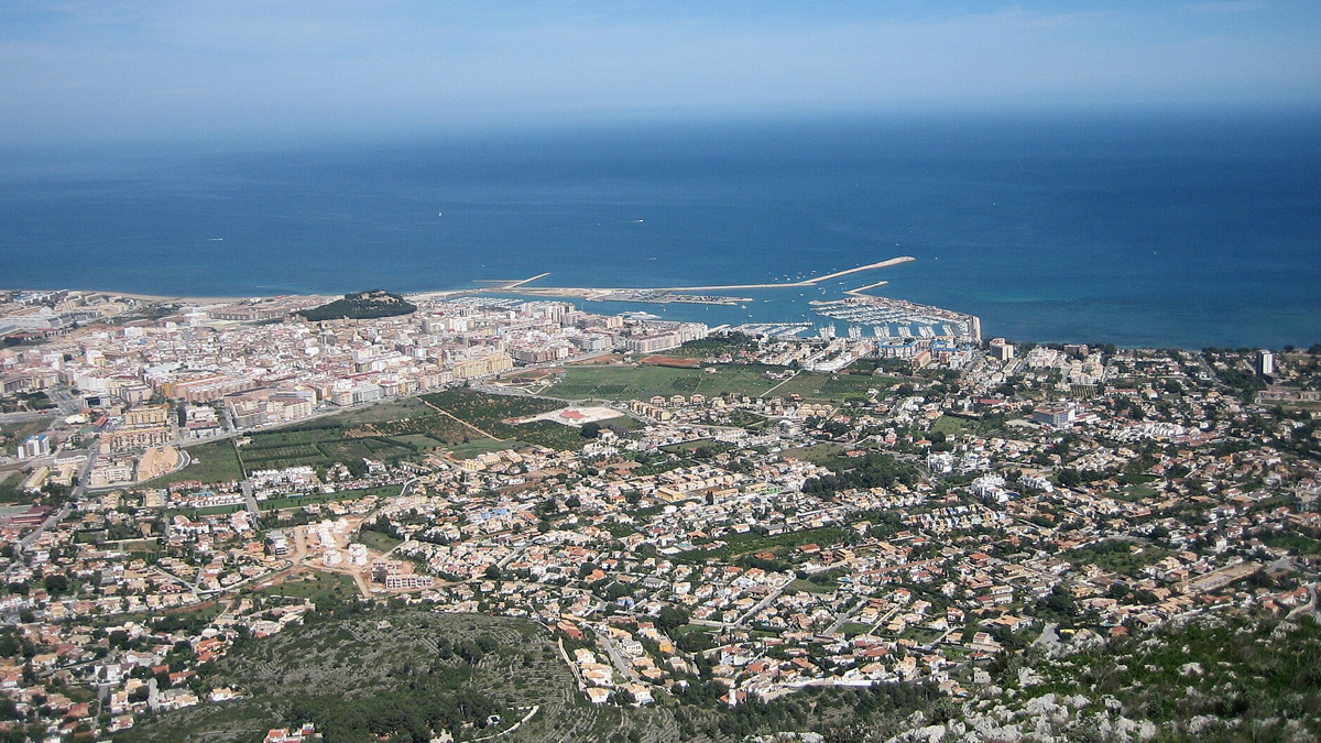 Aerial view of coastal Dénia showing buildings, a harbor, breakwater, and the Mediterranean Sea.