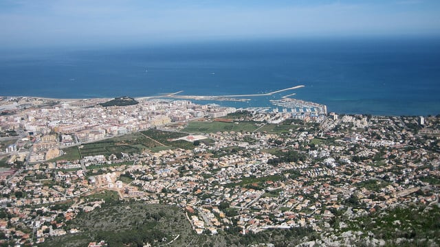 Aerial view of coastal Dénia showing buildings, a harbor, breakwater, and the Mediterranean Sea.