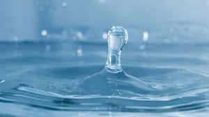 Close-up photograph of a single water droplet splashing and creating ripples on a blue liquid surface
