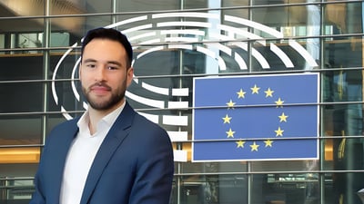 A man in a blue suit stands in front of a glass building featuring the European Parliament logo and the European Union flag.