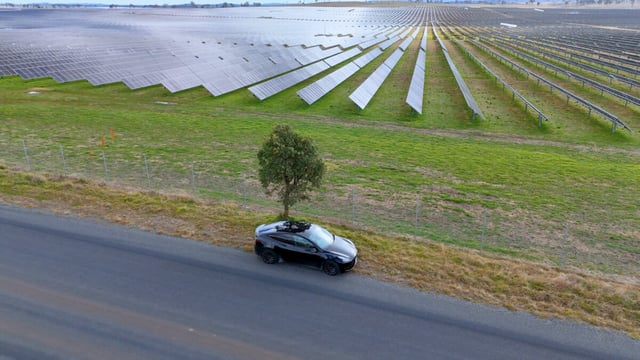 Aerial view of a black electric car parked on a roadside beside a large solar farm with rows of panels stretching across green fields