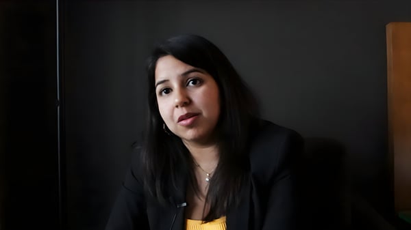 A woman with long dark hair wearing a black blazer and yellow top sits against a dark background.