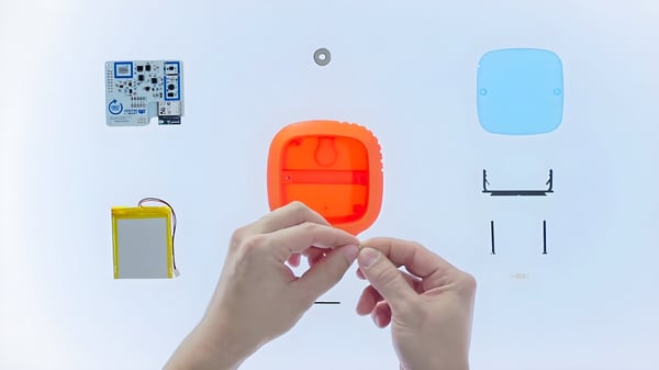 Hands assembling an orange IoT device casing surrounded by a circuit board, battery, and hardware components.