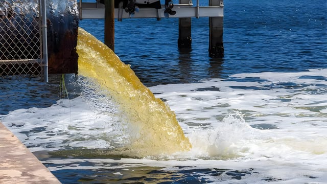 Yellow wastewater discharging from a large metal pipe into a body of water near a pier.