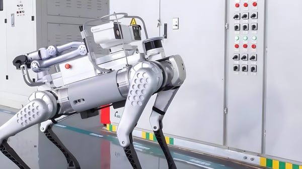 A silver quadrupedal robot stands in an industrial facility facing a control panel with buttons and indicator lights.
